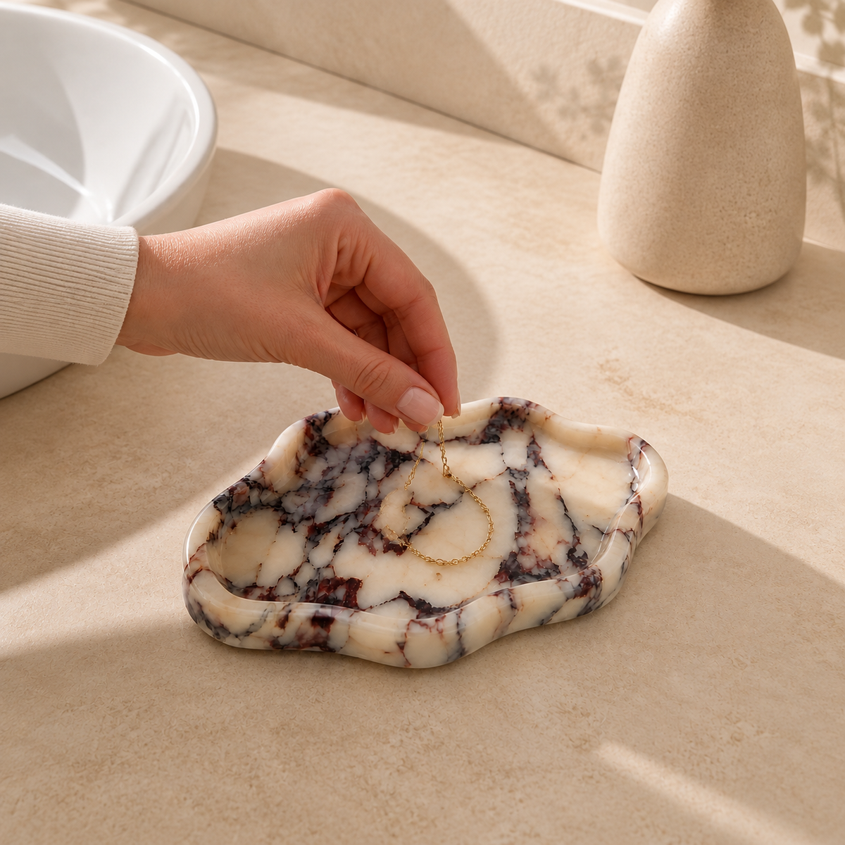 Hand placing a gold necklace on a Calacatta Viola ceramic tray
