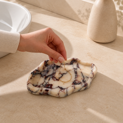 Hand placing a gold necklace on a Calacatta Viola ceramic tray